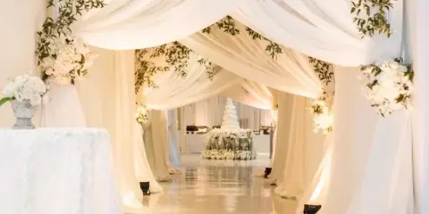 A wedding venue aisle decorated with white draping, greenery, and floral arrangements leading to a wedding cake.