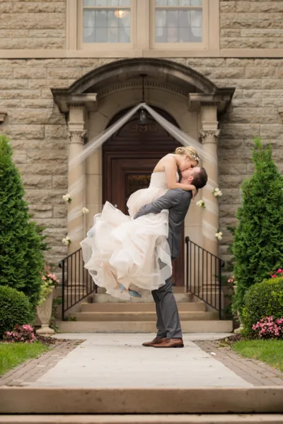 A groom lifts and kisses his bride in front of a stone building entrance.