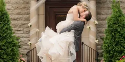 A groom lifts and kisses his bride in front of a stone building entrance.