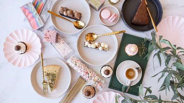 A flat-lay photograph of an assortment of bakery items including cupcakes, cake slices, macarons, and a cup of coffee arranged on a marble surface.