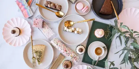 A flat-lay photograph of an assortment of bakery items including cupcakes, cake slices, macarons, and a cup of coffee arranged on a marble surface.
