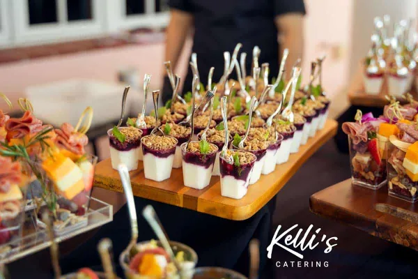 A catering table featuring rows of individual dessert cups and appetizers with a staff member in the background.