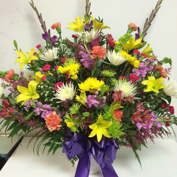 A colorful mixed bouquet of yellow lilies, carnations, and other flowers in a vase with a purple ribbon.