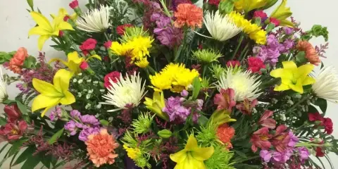 A colorful mixed bouquet of yellow lilies, carnations, and other flowers in a vase with a purple ribbon.