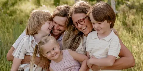A professional outdoor family portrait featuring a smiling couple and three children huddled together in a grassy field.