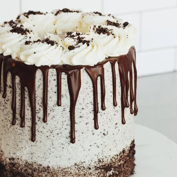 A round cake with white buttercream frosting, a chocolate ganache drip down the sides, and piped cream swirls topped with cookie crumbles.