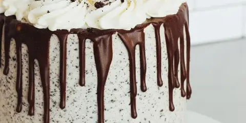 A round cake with white buttercream frosting, a chocolate ganache drip down the sides, and piped cream swirls topped with cookie crumbles.