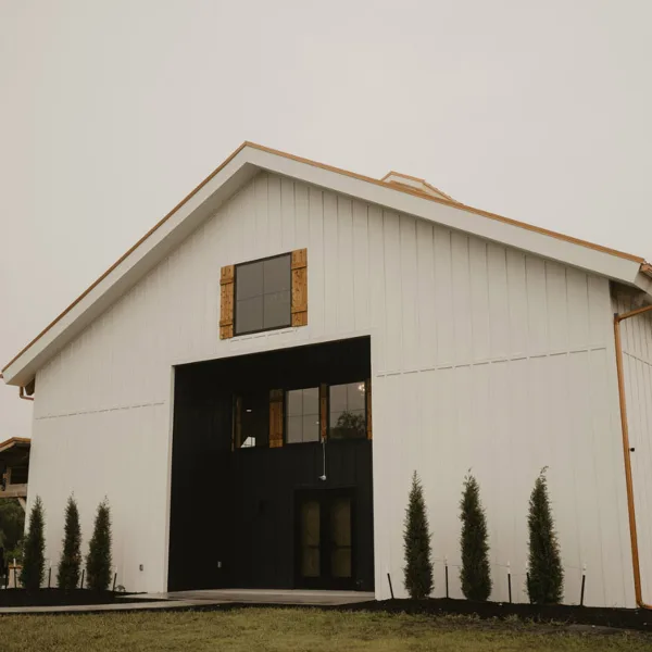 Exterior view of a white barn-style wedding venue with a dark entryway and evergreen trees.