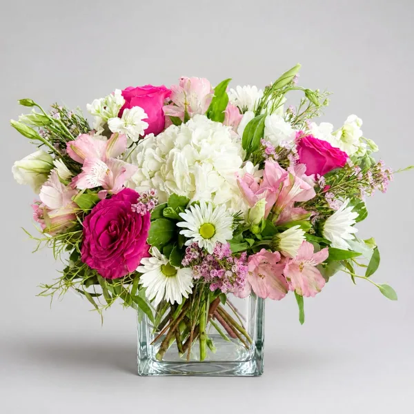A mixed floral arrangement featuring pink and white roses, alstroemeria, and daisies in a clear square glass vase.