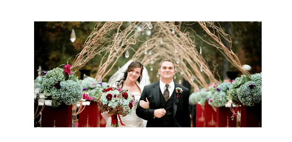 A bride and groom standing in front of a large floral wedding arch with decorated tables on either side.