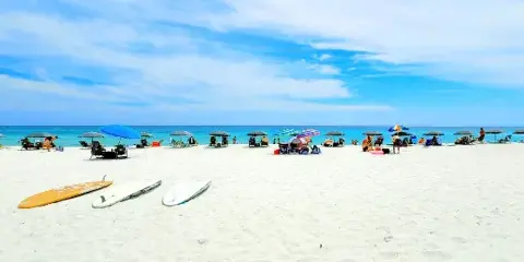 A wide shot of a white sand beach with colorful umbrellas, lounge chairs, and surfboards near the turquoise water.