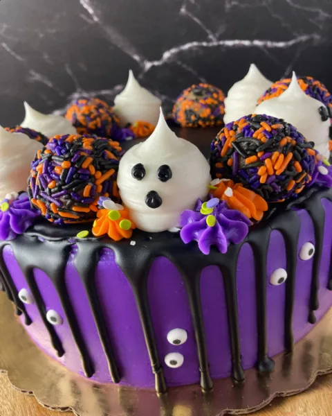 A purple frosted cake decorated with black chocolate drips, purple and orange sprinkles, and a white ghost face.