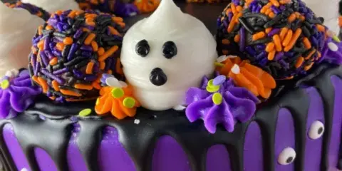 A purple frosted cake decorated with black chocolate drips, purple and orange sprinkles, and a white ghost face.