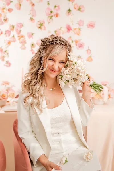 Portrait of a smiling woman in a white suit holding a bouquet of white flowers against a floral wall background.