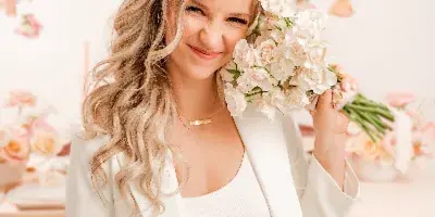 Portrait of a smiling woman in a white suit holding a bouquet of white flowers against a floral wall background.