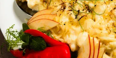 A plated catering dish featuring creamy food, apple slices, red peppers, and herbs with a serving spoon.