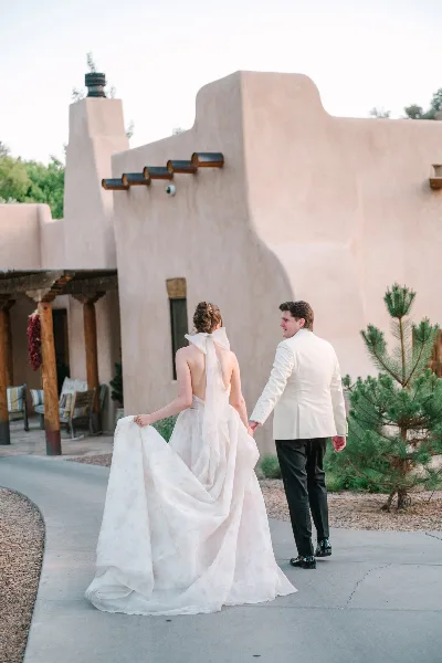 A bride and groom walking hand-in-hand in front of a Santa Fe-style adobe building.