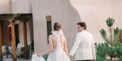 A bride and groom walking hand-in-hand in front of a Santa Fe-style adobe building.