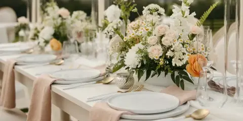 A professionally styled event table featuring white plates, gold cutlery, beige napkins, and a lush floral centerpiece with white and peach roses.