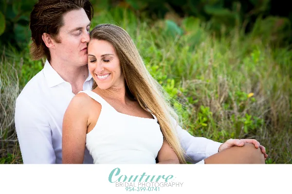 A professional outdoor portrait of a smiling couple in white clothing sitting in a grassy field.