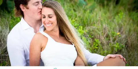 A professional outdoor portrait of a smiling couple in white clothing sitting in a grassy field.