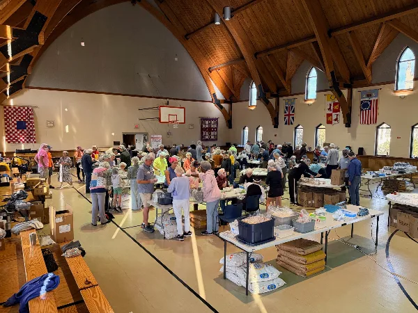 Interior of a large church hall with wooden vaulted ceiling, arched windows, and a basketball hoop, filled with people and tables during a community event.