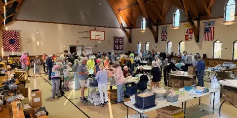 Interior of a large church hall with wooden vaulted ceiling, arched windows, and a basketball hoop, filled with people and tables during a community event.