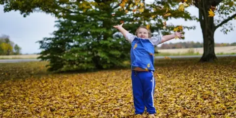 A young child in a blue shirt and pants stands in a field of autumn leaves with arms outstretched, tossing leaves into the air.
