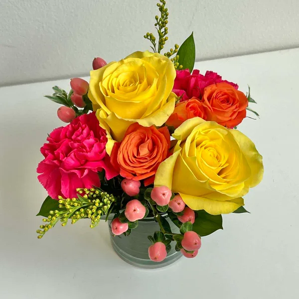 A floral arrangement with yellow and orange roses, pink carnations, and pink berries in a glass vase.