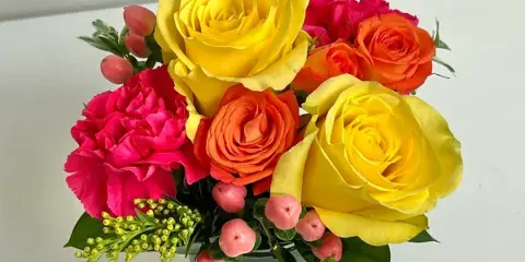 A floral arrangement with yellow and orange roses, pink carnations, and pink berries in a glass vase.