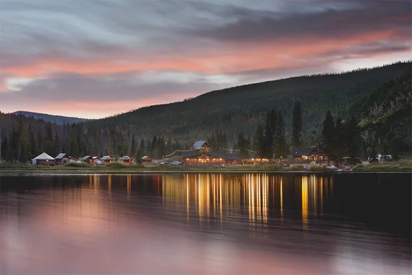 Lakeside view of Piney River Ranch cabins and dining area at dusk with string lights reflecting on the water, surrounded by forested mountains.