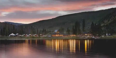 Lakeside view of Piney River Ranch cabins and dining area at dusk with string lights reflecting on the water, surrounded by forested mountains.