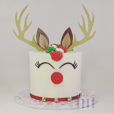 A white specialty cake decorated with fondant to resemble Rudolph the Reindeer, featuring gold antlers, a red nose, and a red collar.