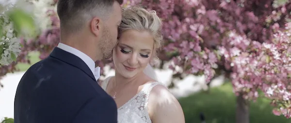 A groom in a dark suit kisses the forehead of a smiling bride in a white lace wedding dress, framed by pink blossoms in a cinematic wedding video still.