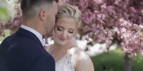 A groom in a dark suit kisses the forehead of a smiling bride in a white lace wedding dress, framed by pink blossoms in a cinematic wedding video still.