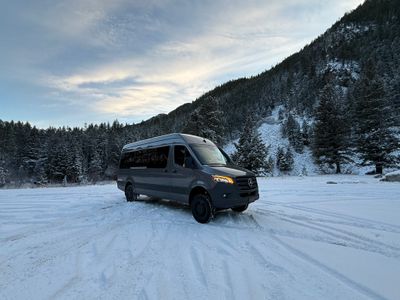 A dark grey luxury Sprinter van parked on a snowy field with a forested mountain in the background.