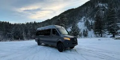 A dark grey luxury Sprinter van parked on a snowy field with a forested mountain in the background.