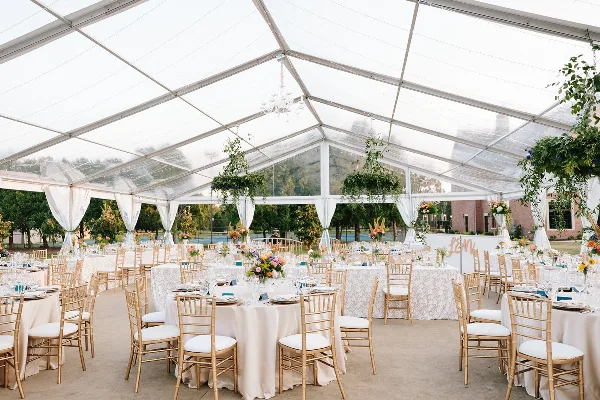 Interior of a clear tent wedding reception featuring round tables with white linens, gold chiavari chairs, and floral centerpieces.
