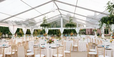 Interior of a clear tent wedding reception featuring round tables with white linens, gold chiavari chairs, and floral centerpieces.