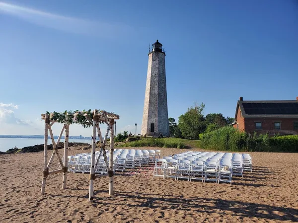 A wedding ceremony setup with a rustic wooden arbor and rows of white chairs arranged on a sandy beach in front of a lighthouse.