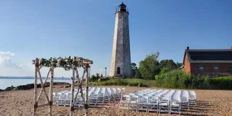 A wedding ceremony setup with a rustic wooden arbor and rows of white chairs arranged on a sandy beach in front of a lighthouse.