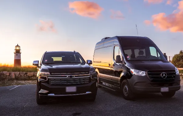 A black Chevrolet SUV and a black Mercedes Sprinter van parked side-by-side at sunset with a lighthouse in the background.