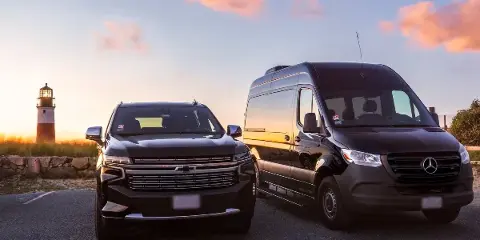 A black Chevrolet SUV and a black Mercedes Sprinter van parked side-by-side at sunset with a lighthouse in the background.