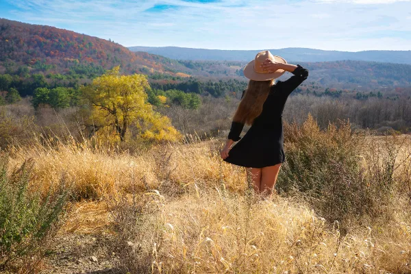 A woman in a black dress and straw hat stands in a field of tall golden grass, looking out over a scenic valley with autumn foliage.