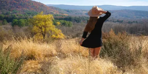 A woman in a black dress and straw hat stands in a field of tall golden grass, looking out over a scenic valley with autumn foliage.