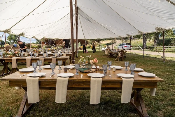 An outdoor wedding reception setup under a white tent with wooden tables, white plates, and floral centerpieces.