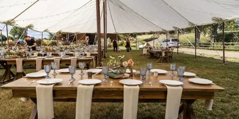 An outdoor wedding reception setup under a white tent with wooden tables, white plates, and floral centerpieces.
