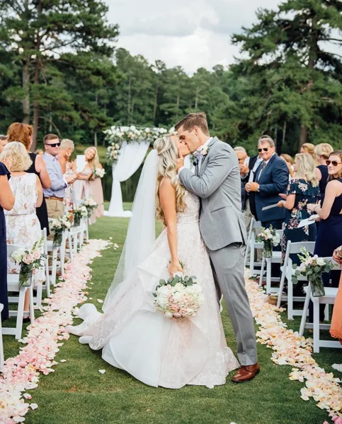 Bride and groom kissing on a grassy aisle lined with flower petals during an outdoor wedding ceremony.