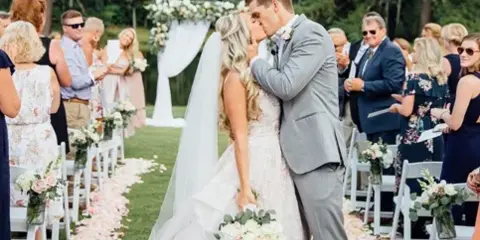 Bride and groom kissing on a grassy aisle lined with flower petals during an outdoor wedding ceremony.