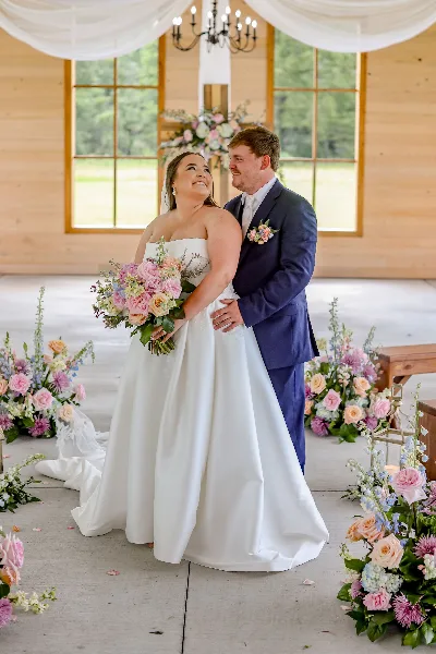 A bride and groom standing together in a rustic wooden venue with floral arrangements and white drapery.